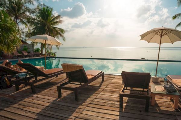 A serene poolside scene with lounge chairs, umbrellas, and a view of the calm sea, set against a backdrop of scattered clouds and palm trees.