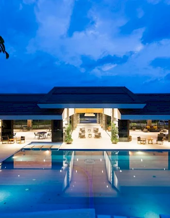 An illuminated outdoor swimming pool area with a modern building in the background, photographed during the evening.