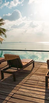 The image shows a serene poolside scene with lounge chairs, umbrellas, palm trees, and a view of the ocean under a partly cloudy sky.