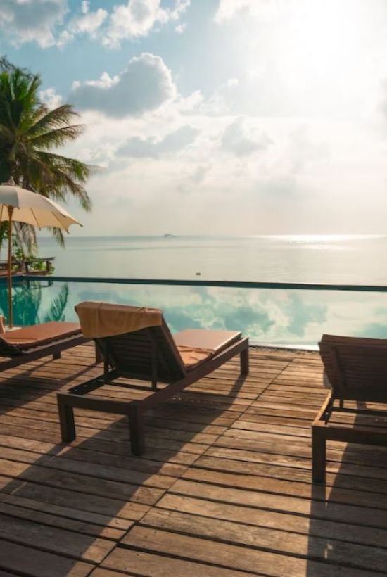 A serene poolside scene with lounge chairs, umbrellas, palm trees, and blue sky reflecting on the calm water, creating a tranquil atmosphere.