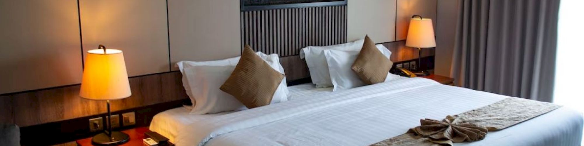 A cozy, well-lit hotel room with a large bed, two bedside tables with lamps, decorative pillows, and a wall-mounted fireplace, all in a modern style.