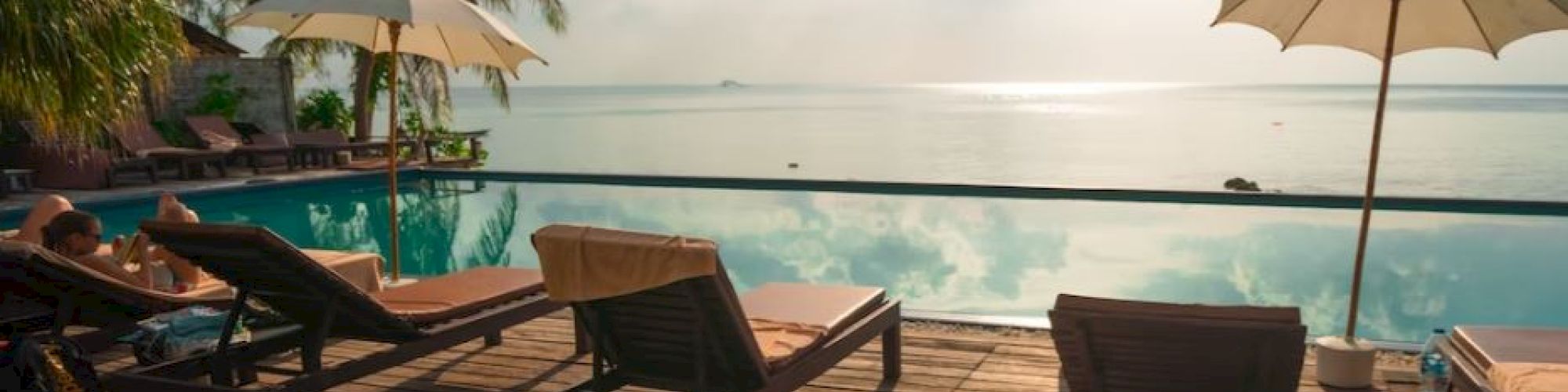The image shows a relaxing poolside scene with lounge chairs, umbrellas, and a calm body of water reflecting the sky.