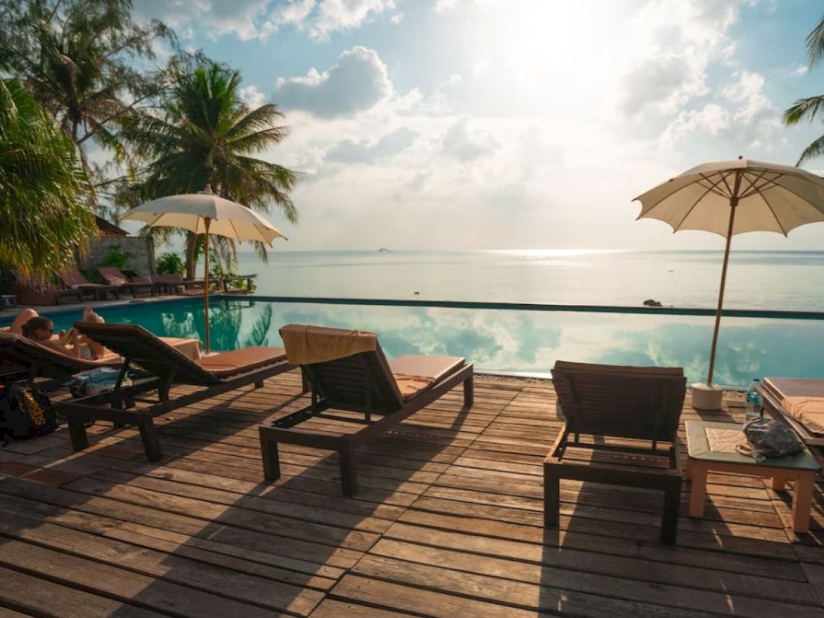 Several lounge chairs and umbrellas are set up by a wooden deck overlooking a serene infinity pool with palm trees and an ocean view.