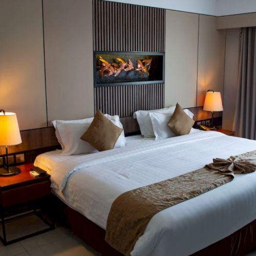 A cozy hotel room with a neatly made bed, decorative pillows, bedside lamps, a wall-mounted fireplace, and a window covered by curtains.