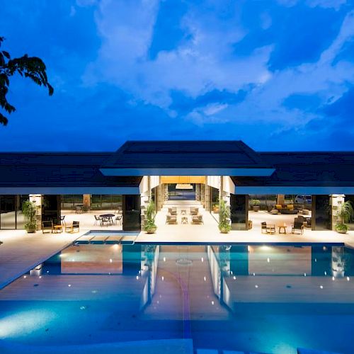 A modern structure with an illuminated pool in the foreground, surrounded by trees and outdoor seating under a dusk sky.