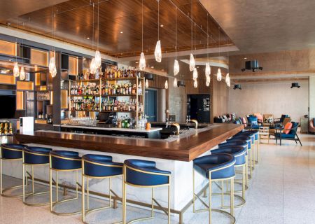 This image shows a modern bar with blue and gold barstools, a fully stocked bar, hanging lights, and an open seating area with tables and chairs.