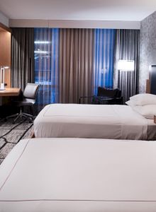 A modern hotel room with two beds, a wall-mounted TV, a desk and chair, and a large window with curtains.