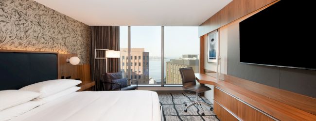 A modern hotel room features a king-sized bed, a wall-mounted TV, a desk with a chair, and a window with an urban view.