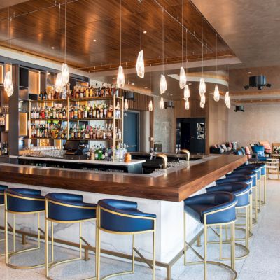 This image depicts a modern bar with a wooden countertop, blue high chairs, and a well-stocked liquor shelf. The ambiance is stylish and inviting.
