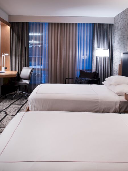 This image shows a modern hotel room with two beds, a wall-mounted TV, a desk with a chair, and stylish lighting and decor.