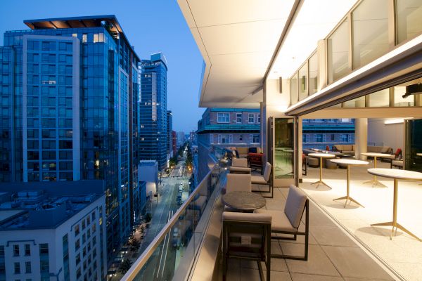 The image shows a modern cityscape at dusk with high-rise buildings alongside a balcony featuring seating arrangements and tables with city views.
