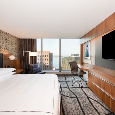 A modern hotel room with a large bed, wall-mounted TV, workspace with chair, large window with city view, and stylish decor.