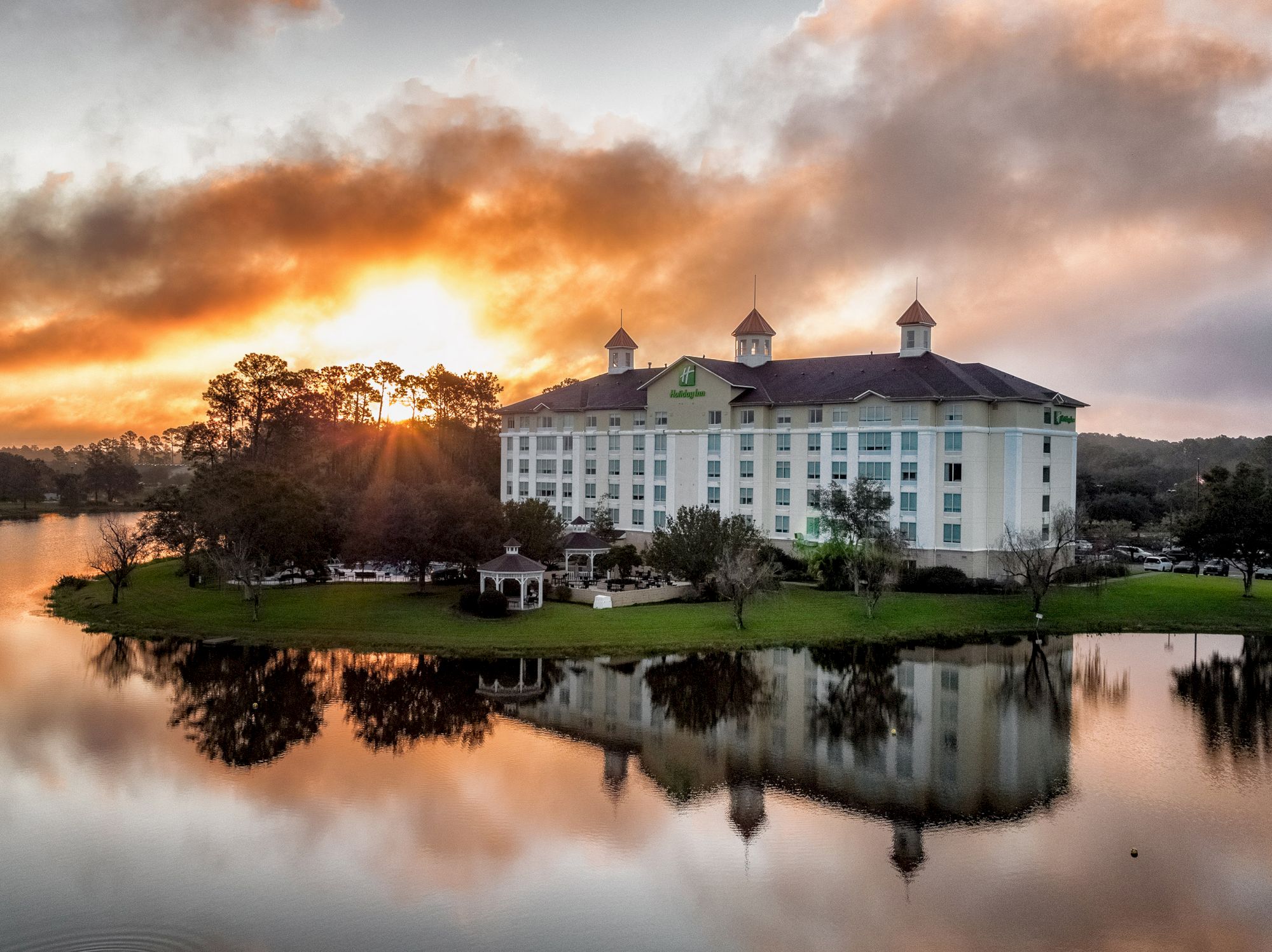 An elegant hotel sits by a serene lake at sunset, with a colorful sky and its reflection mirrored in the water.