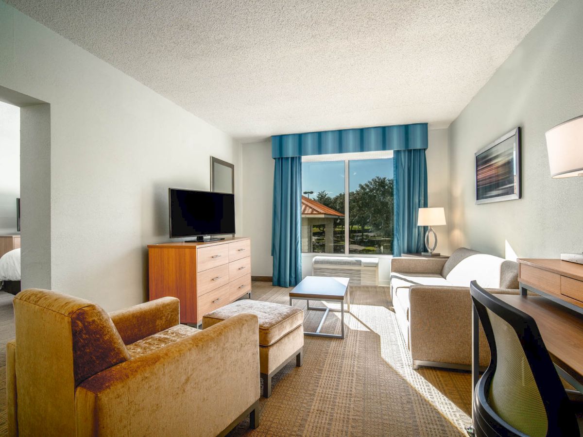 The image shows a modern hotel room with a living area, including chairs, a sofa, TV, desk, and large window with blue curtains overlooking greenery.