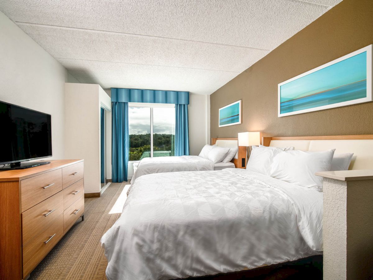 A hotel room with two beds, a dresser with a TV, abstract wall art, a window with blue curtains, and a view of greenery outside.