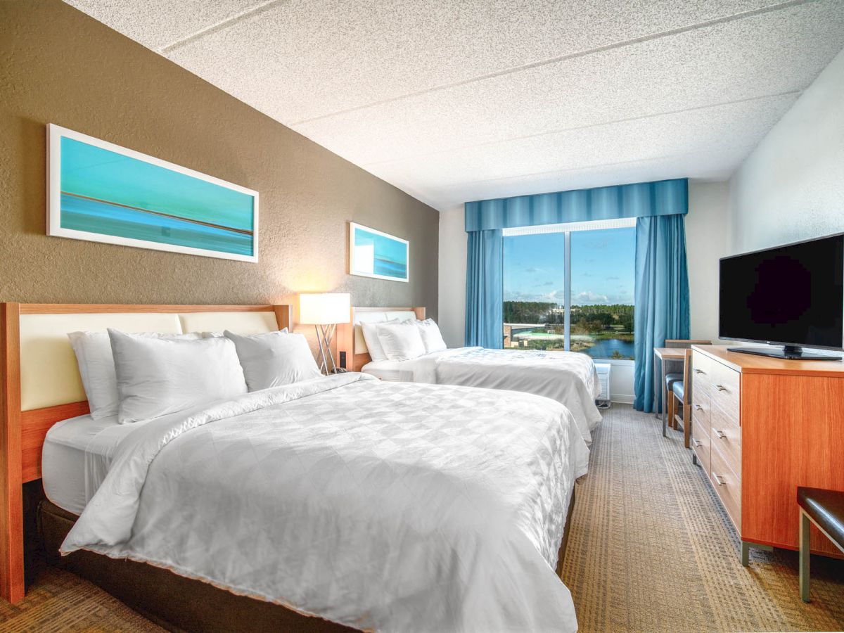 This image shows a hotel room with two beds, a TV, a window with blue curtains, and framed art on the walls. The room appears clean and bright.