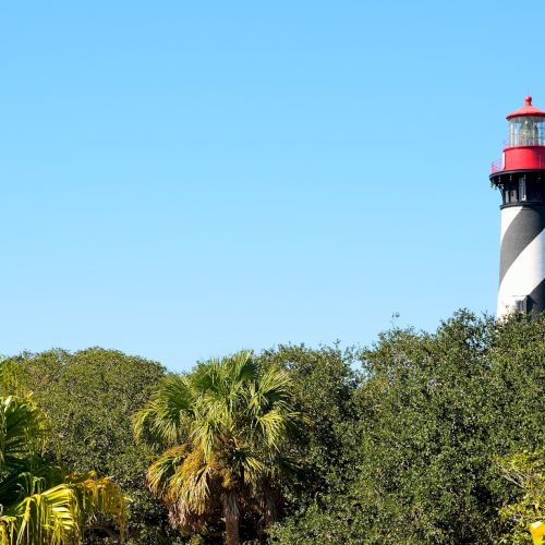 A black-and-white spiral lighthouse with a red top stands tall above green bushes and trees, set against a clear blue sky.