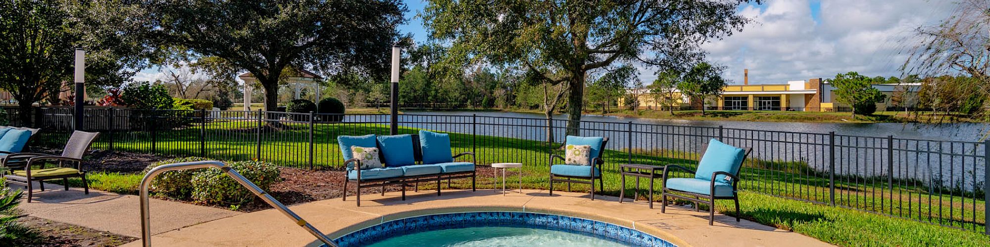 A serene outdoor setting features a small, circular pool bordered by a patio, lounge chairs, and trees, with a lakeside view under a clear blue sky.