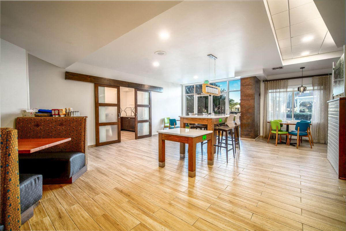 A modern dining area with wooden flooring, various seating options including booths, tables, and bar stools, and a lot of natural light.