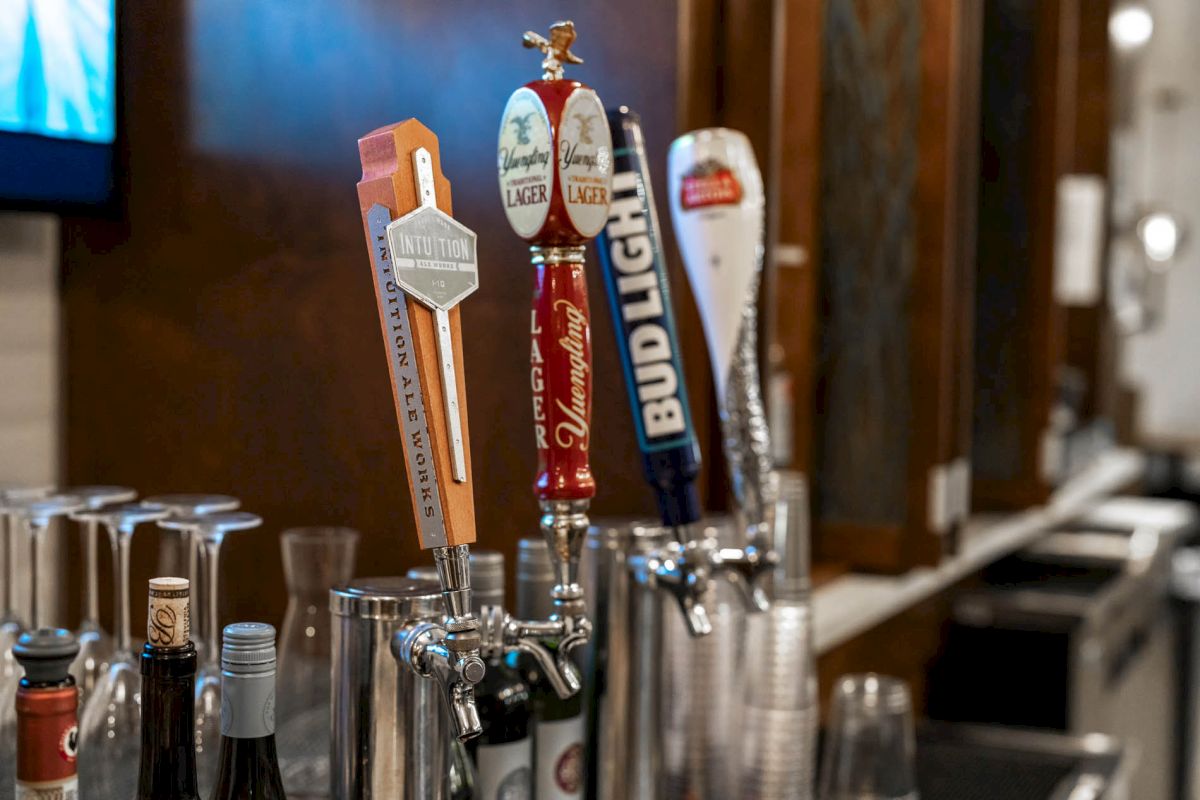 The image shows a bar with four beer taps, featuring various brands, alongside wine bottles and bar equipment in the background.