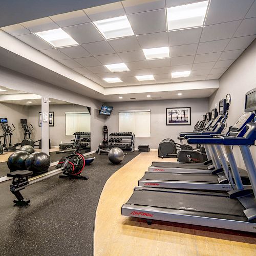 The image shows a modern gym with treadmills, exercise bikes, yoga balls, and weight racks. The room has mirrors, lights, and various equipment.