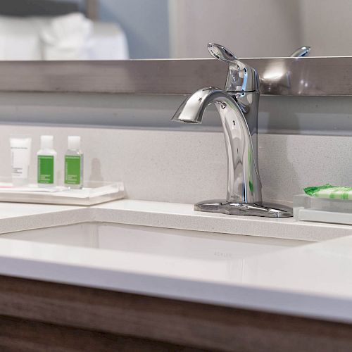 A bathroom sink with a chrome faucet, a mirror, and toiletries including bottles and wrapped soap placed on a white countertop.