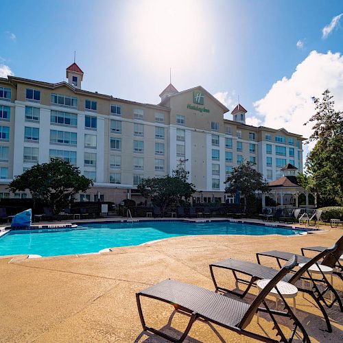 A poolside view of a tall hotel building with multiple floors, surrounded by lounge chairs and trees, under a sunny, blue sky with clouds.