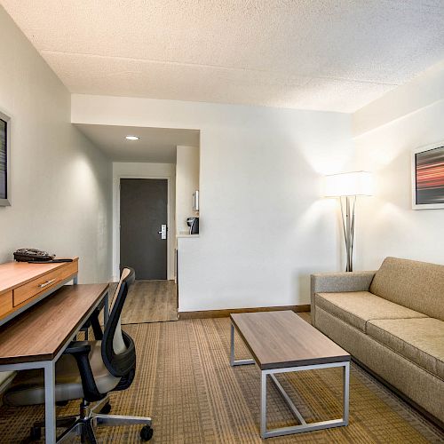 The image shows a modern hotel room with a desk and chair, a lamp, a sofa, a coffee table, and a colorful picture on the wall.