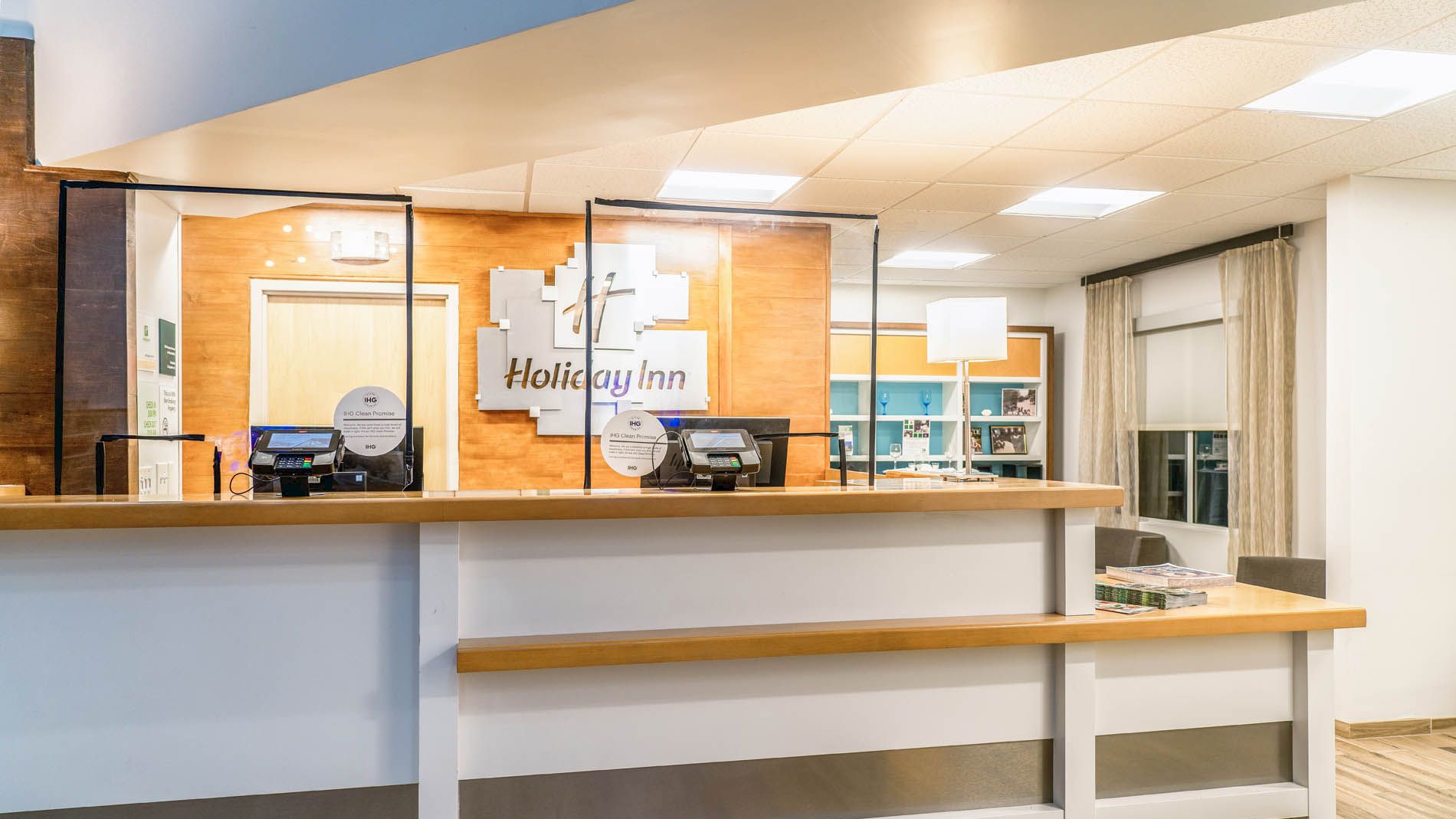 The image shows a hotel reception desk with the "Holiday Inn" logo, computer monitors, and a partition. The area is well-lit with a modern design.