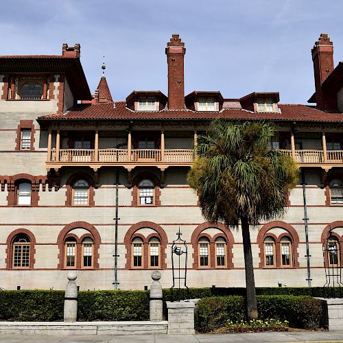 A large, multi-story building with distinct red and cream brickwork, ornate architectural details, and a palm tree out front is shown in the image.