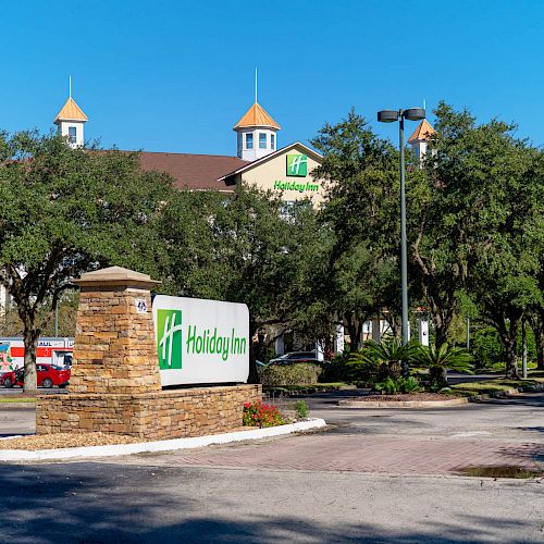 The image shows a Holiday Inn hotel building with a sign in the foreground, surrounded by trees under a clear blue sky.