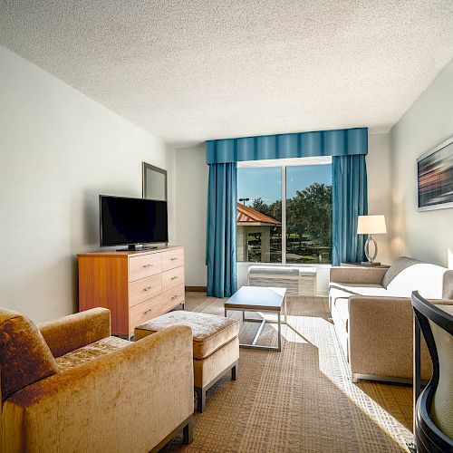 A modern hotel room with seating area, TV, desk, and window with blue drapes; bedroom visible through doorway.