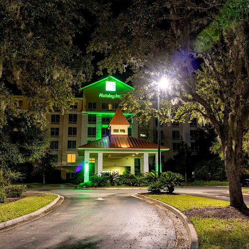 The image shows a well-lit entrance to a Holiday Inn hotel surrounded by trees and a driveway leading to the main entrance of the building.