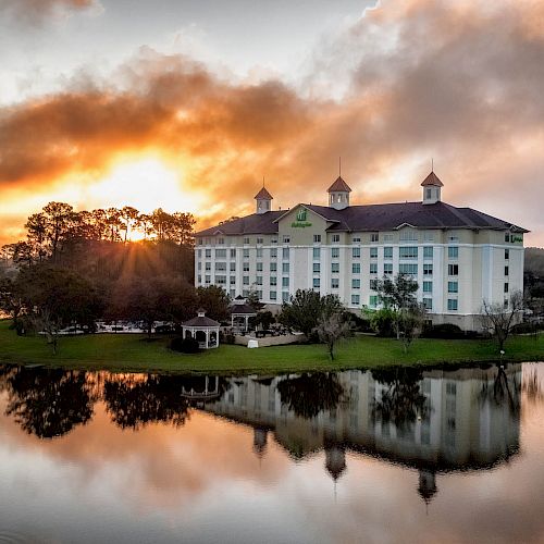 A large building, possibly a hotel, next to a lake with trees and a reflection of the building in the water under a dramatic sky at sunset.