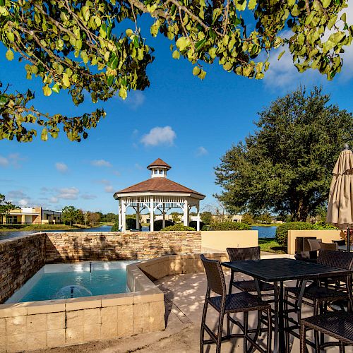 An outdoor patio with wicker furniture, an umbrella, a small pool, and a gazebo by the water under a clear blue sky with trees in the background.