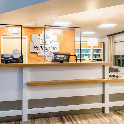 A reception desk in a brightly-lit lobby features protective screens, two computers, and is part of a Holiday Inn as indicated by the sign behind it.