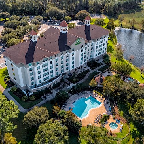 An aerial view of a large hotel with multiple floors, a pool area, surrounding greenery, a lake, and nearby parking lots.