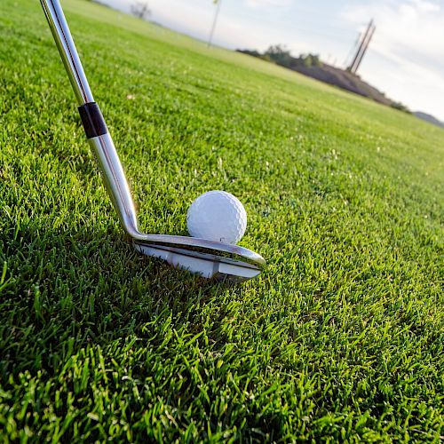 A golf club is positioned behind a golf ball on a lush green fairway, ready for a shot, with another ball visible in the background.