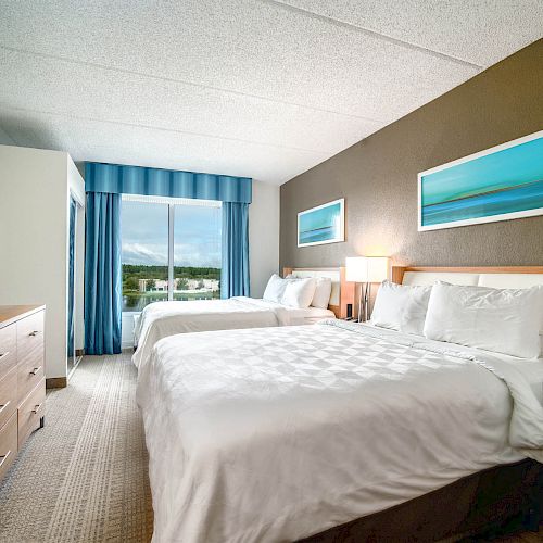 The image shows a hotel room with two large beds, a TV on a wooden dresser, a window with blue curtains, and two framed pictures above the beds.