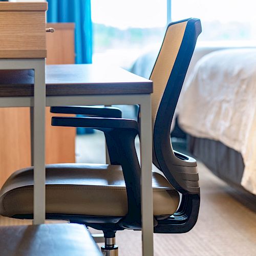 The image shows a side view of an office chair placed at a desk in a room with a bed in the background and blue curtains.