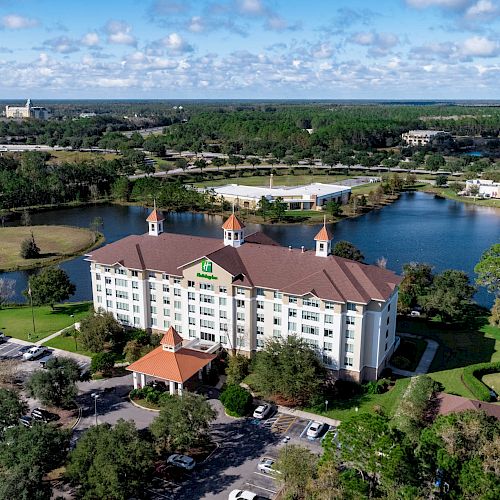 An aerial view of a large hotel building nestled by a lake, surrounded by greenery, with parking areas and roads visible in the background.