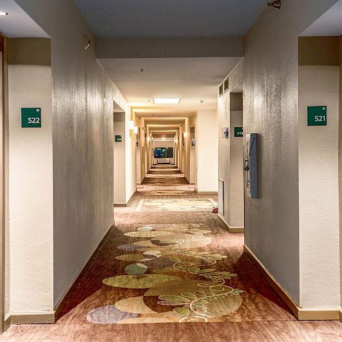 A long, well-lit corridor with doors on both sides in a hotel. Door numbers and room key card locks are visible, and the carpet has a patterned design.