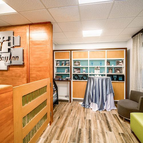 The image shows a Holiday Inn reception area with a wooden counter and a shelving unit behind. There are chairs and a covered table nearby.