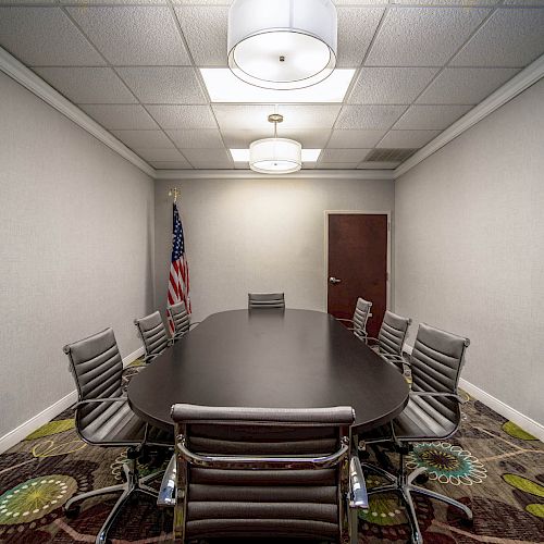 A conference room with a long table, seven chairs, two ceiling lights, and an American flag in the corner, with a door and patterned carpet.