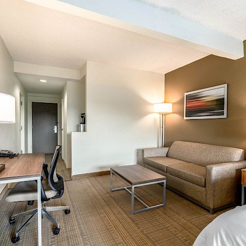 A modern hotel room features a desk and chair, a sofa with side tables, a coffee table, wall art, and lamps, with a bed partially visible.