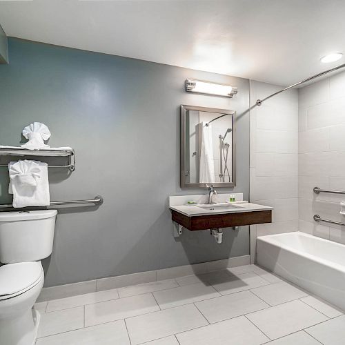 The image shows a clean, modern bathroom with a toilet, sink, mirror, shelves with towels, and a bathtub with a curtain.