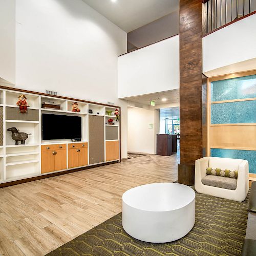 A modern living room with a TV, shelves with décor, a round coffee table, a sofa, and a chair. The space has a high ceiling and wooden floors.