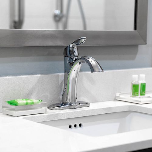 A bathroom sink with a modern faucet, mirror, soap dish, and a tray holding small green bottles and toiletries on a white countertop.