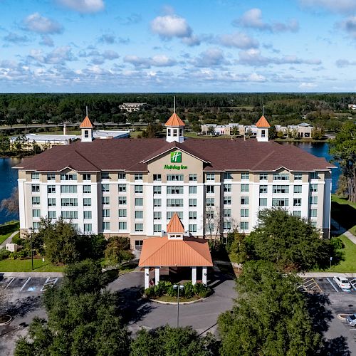 A large hotel with a green logo, surrounded by trees and water, with a parking lot in the foreground and an expanse of forest in the background.