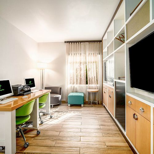 This image shows a modern office space with two desks, computers, shelves, and a large TV screen. There's a window, a lamp, and seating furniture.